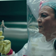 A woman in a hazmat suit holds up infectious fungal tendrils with a pair of tongs.